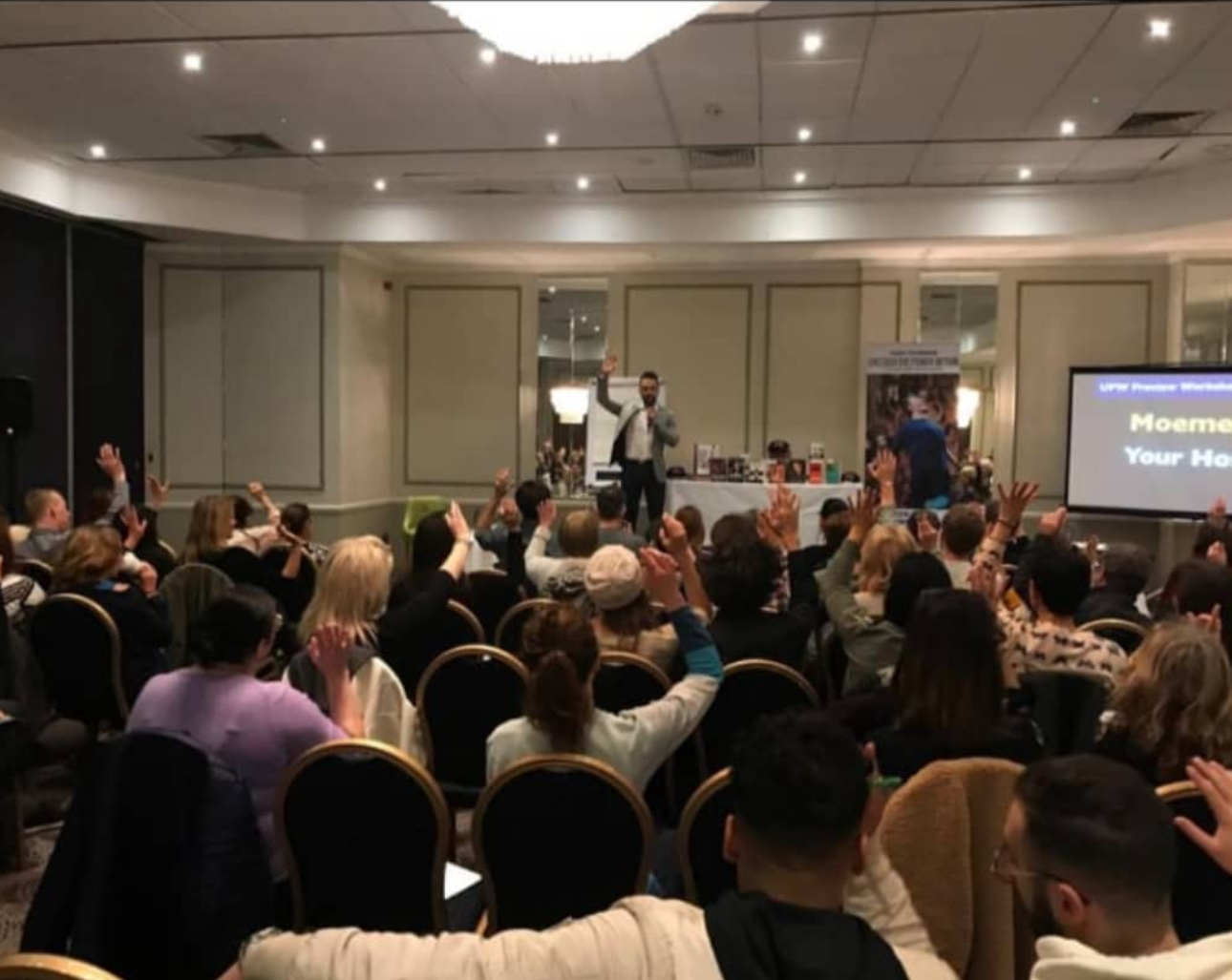 Moemen Lewaa speaking to a room of attendees with their hands raised, with the presentation screen visible.